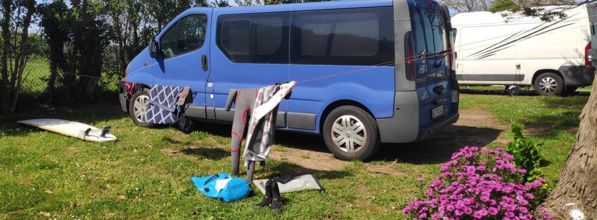 Ein blauer Van, davor eine Wäscheleine mit Wetsuit und Handtüchern. Ein blauer Rucksack und ein Surfboard liegen auf dem Boden