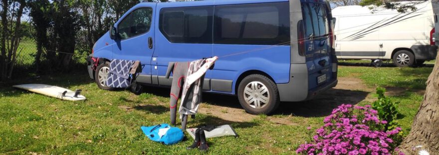 Ein blauer Van, davor eine Wäscheleine mit Wetsuit und Handtüchern. Ein blauer Rucksack und ein Surfboard liegen auf dem Boden